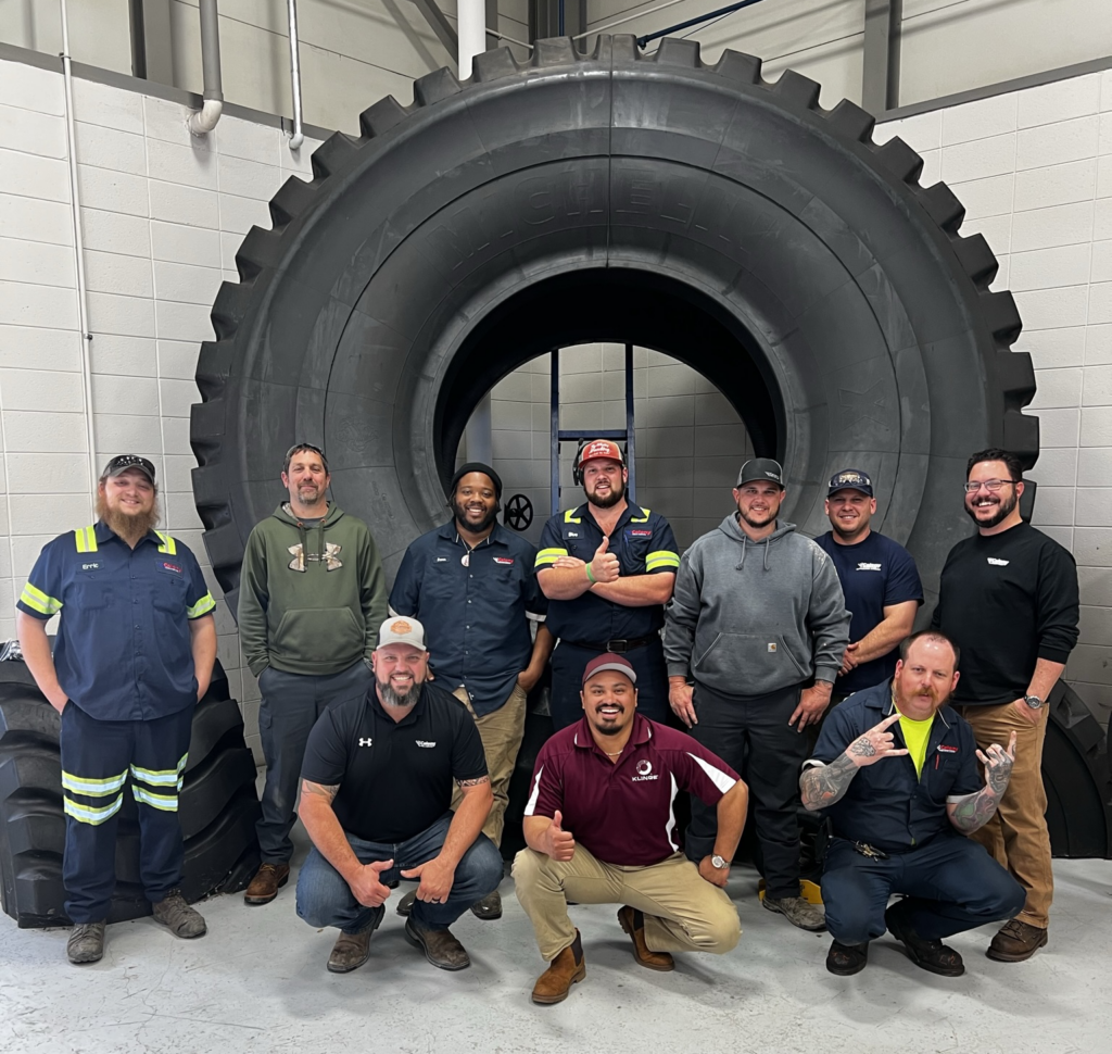 A group of men posing in front of a large off-road tire.