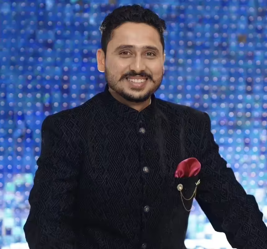 A man in a black suit is smiling against a blue background.